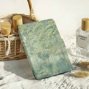 May include: A green and blue e-reader cover with a forest scene. The cover is made of a soft, textured material and has a subtle sheen. The cover is lying on a white, fluffy surface.