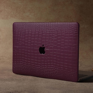 Könnte beinhalten: Eine pflaumenfarbene Laptop-Hülle mit Krokodilhautstruktur. Die Hülle hat einen Ausschnitt für das Apple-Logo. Der Laptop ist geöffnet und zeigt die Tastatur. Ein dekorativer goldener Kerzenhalter befindet sich im Hintergrund.