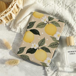 Könnte beinhalten: Ein beiger Notizblock mit einem Zitronen-Druckdesign. Der Notizblock hat weiße Blüten und grüne Blätter. Der Notizblock liegt auf einer weißen Oberfläche mit einem Weidenkorb und einer Parfümflasche.