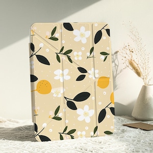 Puede incluir: Una funda beige para iPad con un patrón floral que presenta flores blancas, hojas verdes y limones amarillos. La funda tiene un diseño plegable.
