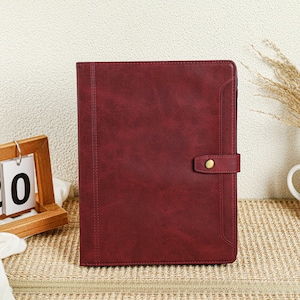 Peut inclure: Un carnet relié en cuir bordeaux avec une fermeture à bouton doré. Le carnet est posé sur un tapis tissé.