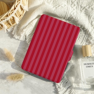 May include: A red and pink striped tablet case rests on a white textured blanket. The case is rectangular with vertical stripes. A wicker basket with a wooden lid and a perfume bottle are in the background.