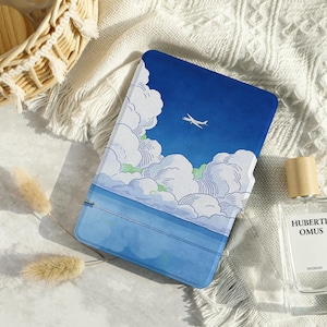 May include: A blue and white notebook with a sky and cloud design, featuring a small airplane. The notebook is rectangular and appears to be closed. The background includes a white textured surface and a perfume bottle.