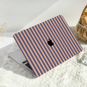 Może przedstawiać: Laptop z obudową w pionowe paski w kolorze granatowym i brzoskwiniowym. Laptop jest otwarty, widoczna jest klawiatura i ekran. Widoczne jest logo Apple. Laptop znajduje się na białej powierzchni.
