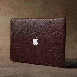 Puede incluir: Una funda marrón para portátil con un patrón texturizado similar a la piel de cocodrilo. La funda presenta el logotipo de Apple en blanco. El portátil está parcialmente abierto, revelando el teclado. Un candelabro dorado decorativo está en el fondo.