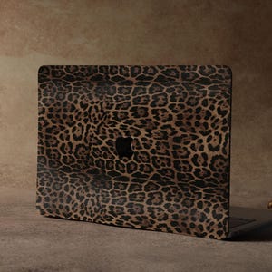 Könnte beinhalten: Ein Laptop mit einem Leopardenmuster-Cover. Das Cover zeigt ein Muster aus braunen und schwarzen Flecken auf einem beigen Hintergrund. Der Laptop ist geöffnet und zeigt das schwarze Apple-Logo. Rechts befindet sich ein goldfarbener Kerzenhalter.