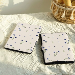 May include: Two white e-reader cases with a floral pattern featuring blue flowers and green stems. The cases are on a white crocheted surface.