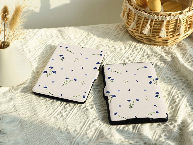 May include: Two white e-reader cases with a floral pattern featuring blue flowers and green stems. The cases are on a white crocheted surface.