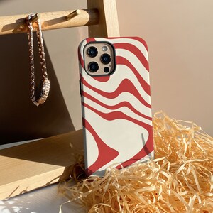 May include: A white phone case with a red abstract swirl pattern. The case is designed for a phone with a triple lens camera system.