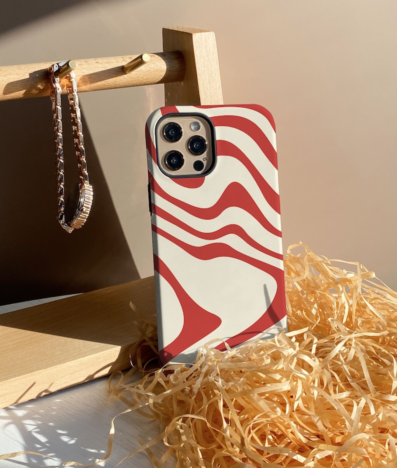 May include: A white phone case with a red abstract swirl pattern. The case is designed for a phone with a triple lens camera system.