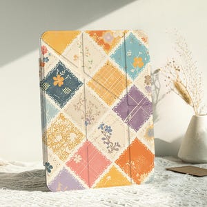 Könnte beinhalten: Eine farbenfrohe Tablet-Hülle mit Patchwork-Design. Die Hülle zeigt rautenförmige Muster in Gelb, Blau, Orange, Rosa und Lila. Jede Raute hat ein einzigartiges Blumen- oder Spitzendesign. Die Hülle steht auf einer weißen Oberfläche.