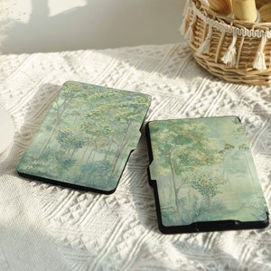 May include: Two black e-reader cases with a forest scene design. The cases have a light green and white background with a watercolor effect. The cases are on a white textured surface.