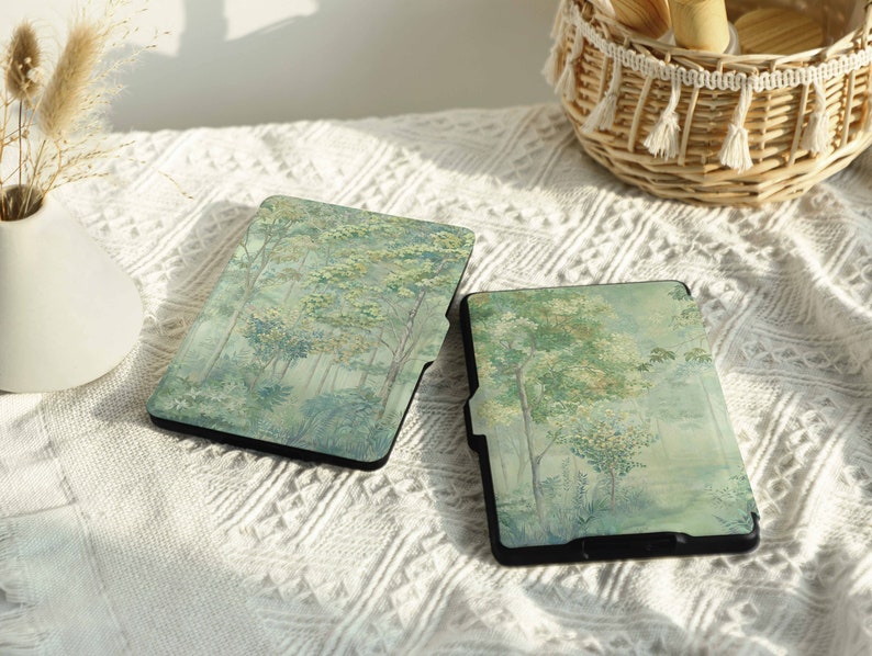 May include: Two black e-reader cases with a forest scene design. The cases have a light green and white background with a watercolor effect. The cases are on a white textured surface.