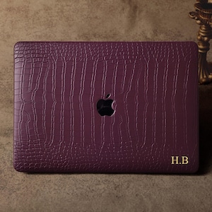 Könnte beinhalten: Eine tiefviolette Laptop-Hülle mit einem strukturierten Krokodilhautmuster. Die Hülle hat eine Aussparung für das Apple-Logo und goldene Initialen "H.B." in der unteren rechten Ecke. Die Hülle ist für einen Laptop konzipiert.