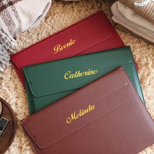 Könnte beinhalten: Drei personalisierte Laptop-Hüllen aus Leder in Rot, Grün und Braun. Jede Hülle hat einen Namen in Gold gestickt: "Beenle", "Catherine" und "Melinda". Die Hüllen sind auf einer strukturierten, hellbraunen Oberfläche angeordnet.