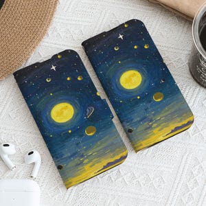 Może przedstawiać: Dwa etui na telefon z motywem gwieździstej nocy. Etui mają ciemnoniebieskie tło z żółtymi księżycami, gwiazdami i planetami. Projekt przypomina obraz Van Gogha. Etui leżą na białej powierzchni.