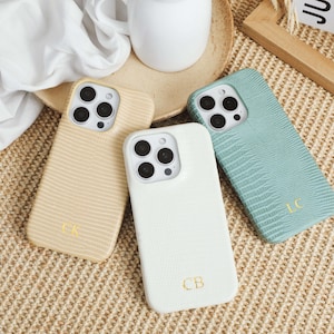 Könnte beinhalten: Drei Smartphone-Hüllen mit einer strukturierten Eidechsenhaut-Optik in Beige, Weiß und Salbeere und Salbeigrün. Jede Hülle verfügt über einen Ausschnitt für die Dreifachkamera und goldfarbene geprägte Initialen: CK auf der beigen, CB auf der weißen und LC auf der grünen Hülle.
