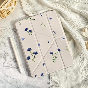 Puede incluir: Una funda para iPad de color crema con un patrón geométrico y detalles florales azules. La funda está sobre una manta de ganchillo blanca.