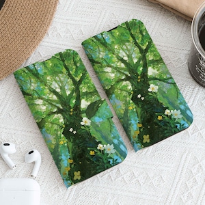 Puede incluir: Dos fundas para teléfono con una vibrante escena de bosque que presenta un gran árbol con hojas verdes, flores blancas y amarillas y un cielo azul. Las fundas están abiertas. También se ven un sombrero de paja, una taza de café y auriculares inalámbricos blancos.