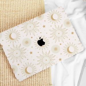 Op de afbeelding: Een laptop met een crèmekleurige hoes met een hemels patroon van zonnen, manen en sterren in goud. Het Apple-logo is zichtbaar in zwart. De laptop ligt op een wit oppervlak, met een smartphone en decoratieve items in de buurt.