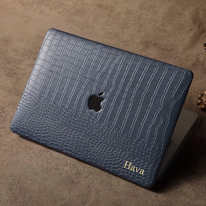 Könnte beinhalten: Eine marineblaue Laptop-Hülle mit Krokodilhaut-Textur. Die Hülle hat einen Ausschnitt für das Apple-Logo und den Namen "Hava" in goldfarbenen Buchstaben. Die Hülle liegt auf einer braunen Oberfläche.