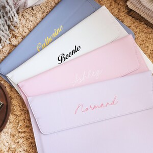 Könnte beinhalten: Eine Sammlung personalisierter Dokumentenmappen in Pastellfarben. Die Mappen sind in Blau, Weiß, Rosa und Lavendel gehalten, jede mit einem Namen in einer kontrastierenden Schriftart. Die Artikel sind auf einer strukturierten, hellbraunen Oberfläche angeordnet.