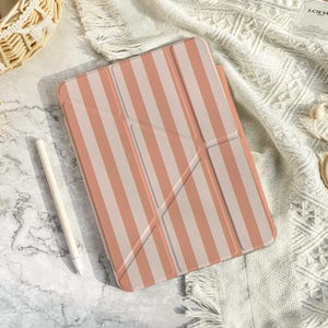 Könnte beinhalten: Eine Tablet-Hülle mit rosa-weißem Streifenmuster. Ein weißer Stift liegt neben der Hülle. Die Hülle befindet sich auf einer weißen, strukturierten Oberfläche. Ein Weidenkorb und ein Holzspielzeug sind ebenfalls im Bild.