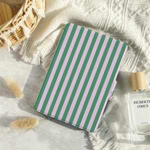 Könnte beinhalten: Ein grün-rosa gestreifter Notizbuch- oder Tablet-Cover liegt auf einer weißen, strukturierten Oberfläche. Der Cover hat vertikale Streifen. Eine Parfümflasche und ein Weidenkorb befinden sich im Hintergrund.
