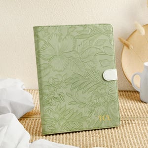 Puede incluir: Un diario de cuero verde con un diseño floral en relieve. El diario tiene una solapa de cierre de cuero blanco y las iniciales "KCA" están grabadas en la portada.