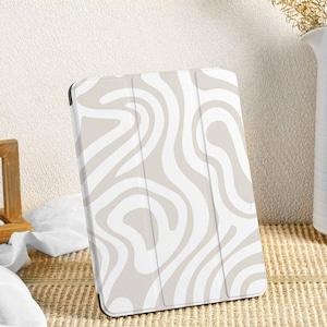 Peut inclure: Étui pour tablette blanc avec un motif abstrait en spirale beige. L'étui est fermé et debout sur une surface tissée.