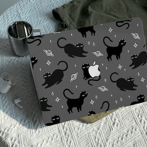 Könnte beinhalten: Eine graue Laptop-Haut mit einem sich wiederholenden Muster aus schwarzen Katzen und weißen Sternen, Planeten und Symbolen. Die Haut hat eine matte Oberfläche.