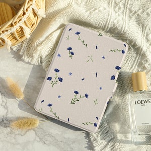 May include: A beige floral patterned e-reader case with blue flowers and green stems. The case is lying on a white surface with a wicker basket and a bottle of perfume.