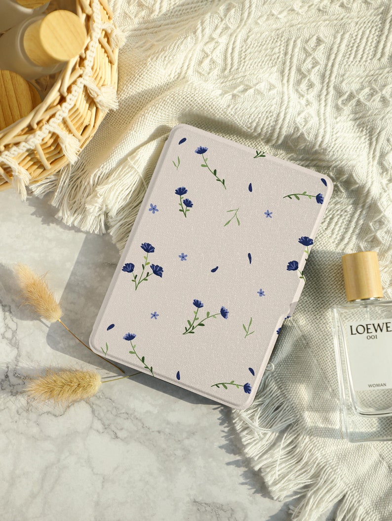 May include: A beige floral patterned e-reader case with blue flowers and green stems. The case is lying on a white surface with a wicker basket and a bottle of perfume.