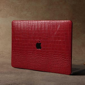 Könnte beinhalten: Eine rote Laptop-Hülle mit Krokodilhaut-Textur. Die Hülle hat einen Ausschnitt für das Apple-Logo. Der Laptop befindet sich auf einem neutralen Hintergrund.