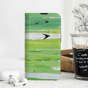 Pode incluir: Capa de telefone com arte abstrata verde e branca com três pássaros pretos em voo. Ao lado da capa, um copo de líquido escuro com texto em francês. Fones de ouvido sem fio brancos e estojo de carregamento em primeiro plano.