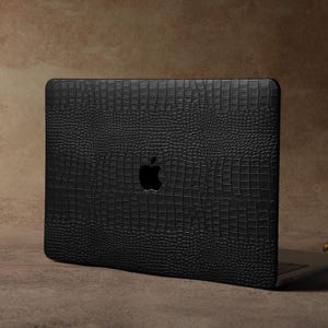 Könnte beinhalten: Schwarze Laptop-Hülle mit Krokodilhaut-Textur. Das Gehäuse hat das Apple-Logo und befindet sich auf einer braunen Oberfläche. Rechts vom Laptop befindet sich ein goldfarbener dekorativer Kerzenhalter.