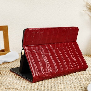 May include: A red leather tablet case with a crocodile pattern. The case is closed and has a black flap.