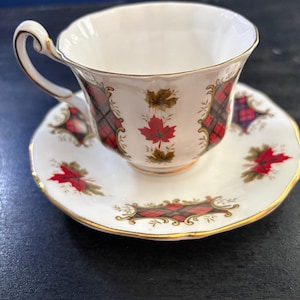 May include: White teacup and saucer set with gold trim. The teacup and saucer feature a red and black plaid pattern with red maple leaf accents. The teacup has a curved handle and the saucer has a scalloped edge.