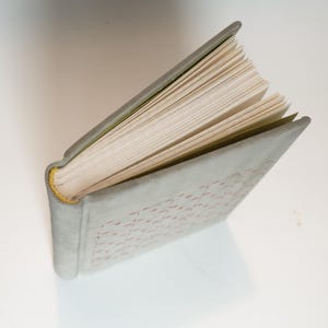 May include: An open book with a light gray cover and a yellow spine. The pages are off-white and slightly fanned out. The cover has a subtle pattern. The book is angled on a white surface.