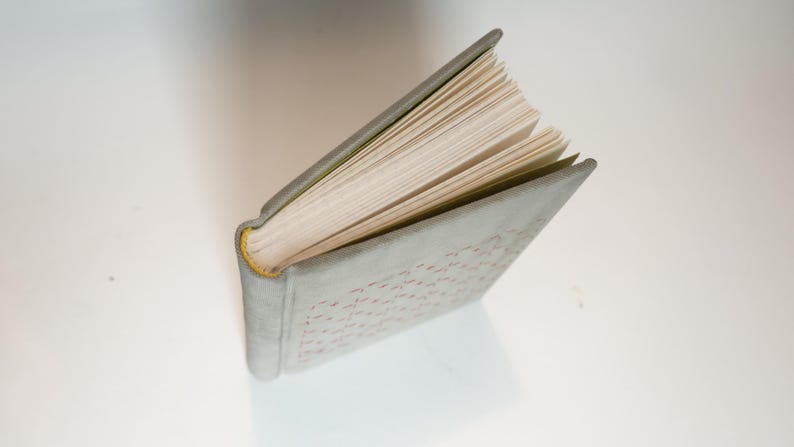 May include: An open book with a light gray cover and a yellow spine. The pages are off-white and slightly fanned out. The cover has a subtle pattern. The book is angled on a white surface.