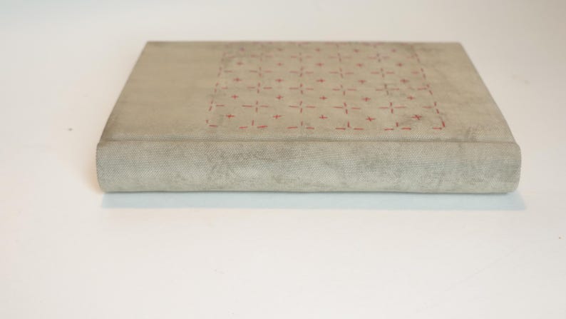 May include: A rectangular, light gray book with a textured cover. The top surface features a grid pattern of small red cross-like designs. The spine is rounded, and the book appears to be closed.
