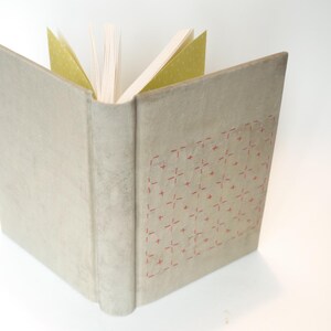 May include: An open book with a light beige cover and a spine. The cover has a stitched pattern in red. The pages are white, with some gold-colored pages visible. The book is set against a white background.
