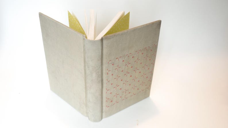 May include: An open book with a light beige cover and a spine. The cover has a stitched pattern in red. The pages are white, with some gold-colored pages visible. The book is set against a white background.