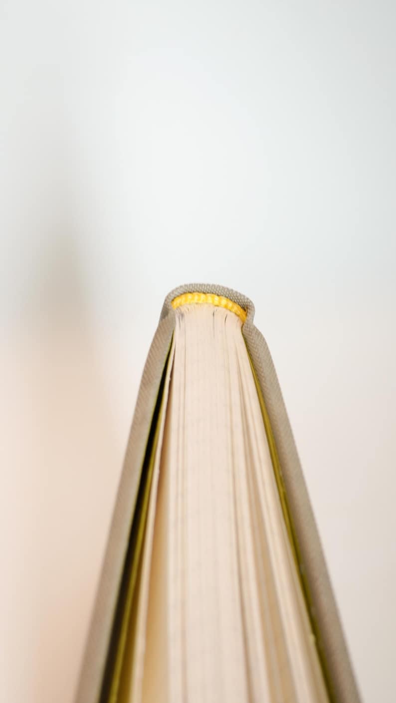May include: Close-up of a book's spine and pages. The book has a gray cover with a yellow top edge. The pages are off-white, and the book is set against a plain white background.