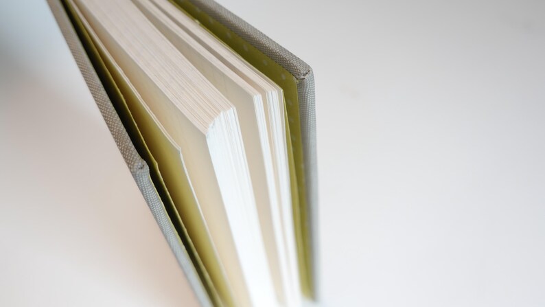 May include: Close-up of a hardcover book with a textured gray spine and light-colored pages. The pages are slightly fanned out, revealing their edges. The book's cover is a muted green color.