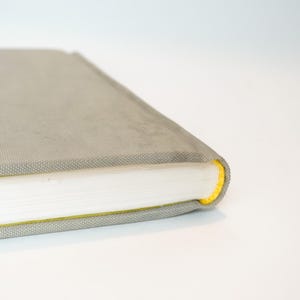 May include: A gray book with a textured cover and white pages. The edge of the book is accented with a bright yellow color. The book is closed and resting on a white surface.