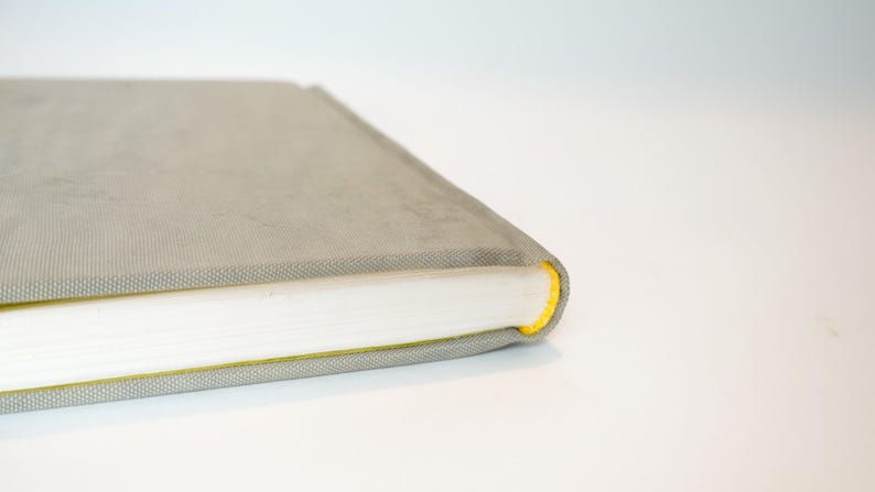 May include: A gray book with a textured cover and white pages. The edge of the book is accented with a bright yellow color. The book is closed and resting on a white surface.