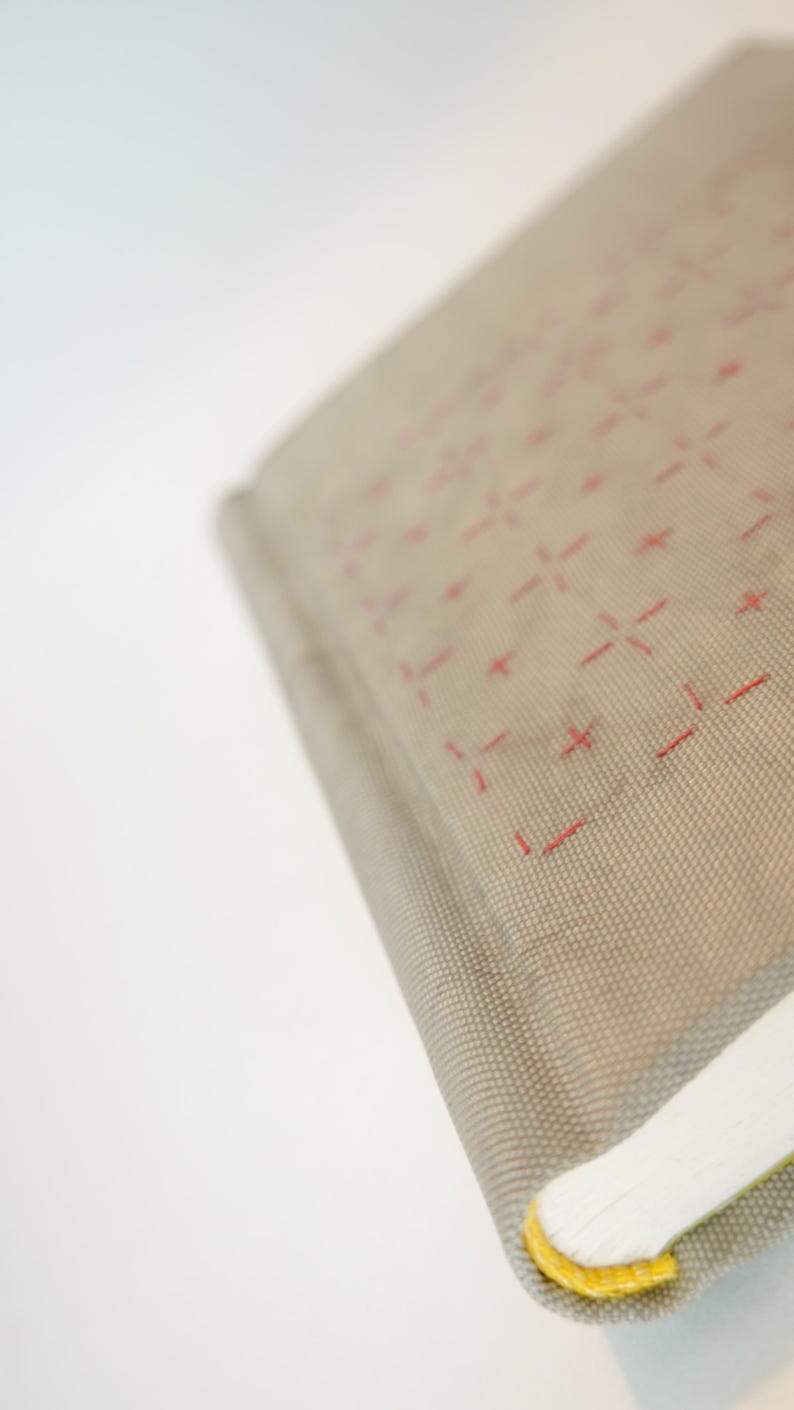 May include: Close-up of a book with a light gray cover featuring a grid pattern of red dashes. The book's pages are white, and the spine has a yellow accent.