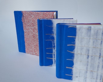 May include: Three handmade journals with blue covers and different decorative spines. The first journal has a red and white floral pattern, the second has a white fabric spine, and the third has a white fabric spine with a woven pattern.