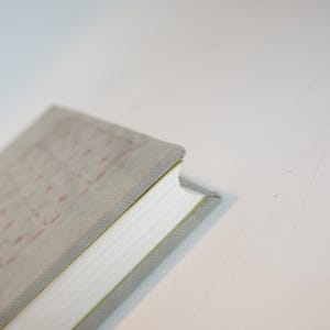 May include: A light gray book with a textured cover and white pages. The cover has a subtle grid pattern in red. The book's spine is visible, showing the edge of the pages. The book is angled, with a plain white background.